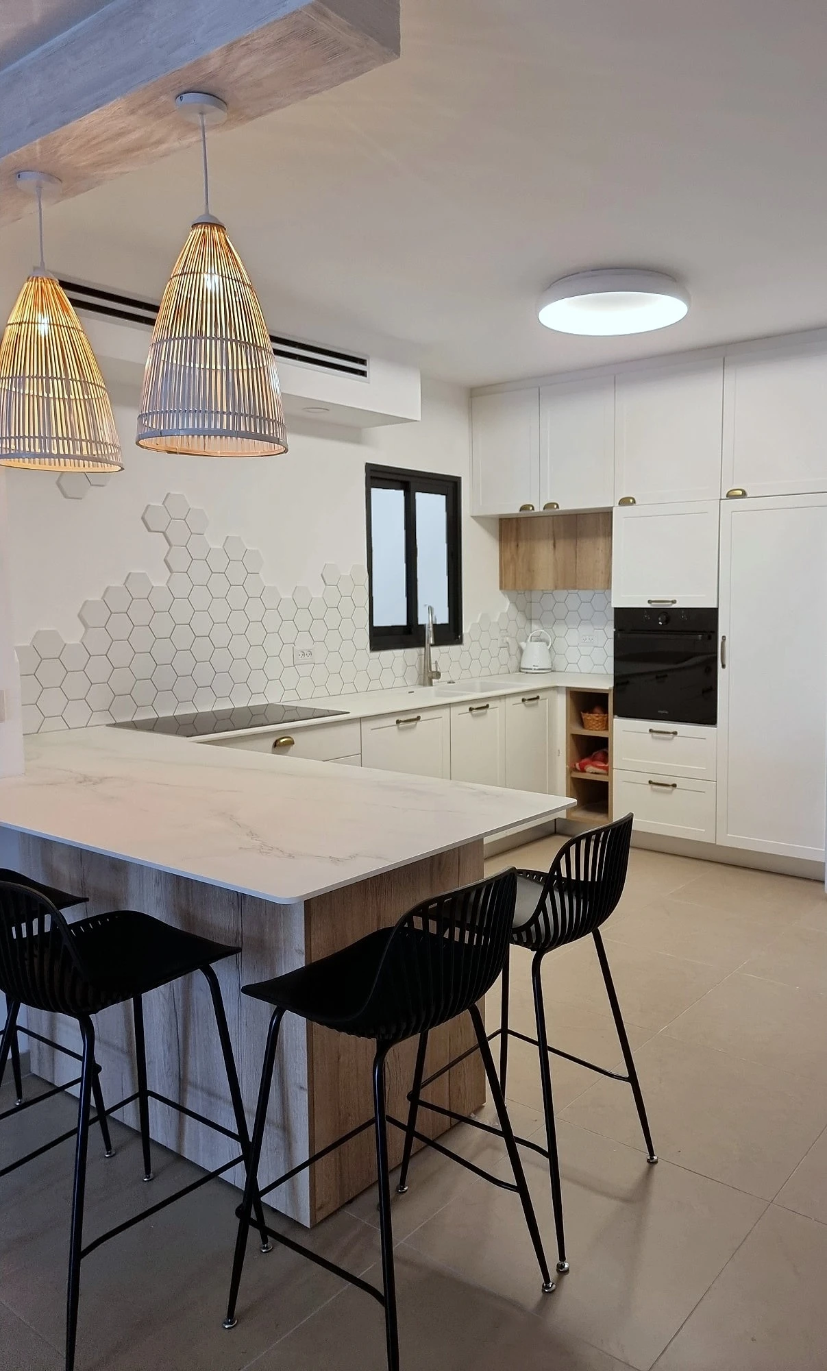 חצי אי עם משטח שיש וכסאות בר שחורים, גופי תאורה תלויים מנצרים ומטבח לבן מאחור